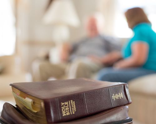 A Bible on top of a scripture case on a table, with a man and woman sitting on a couch in the background.