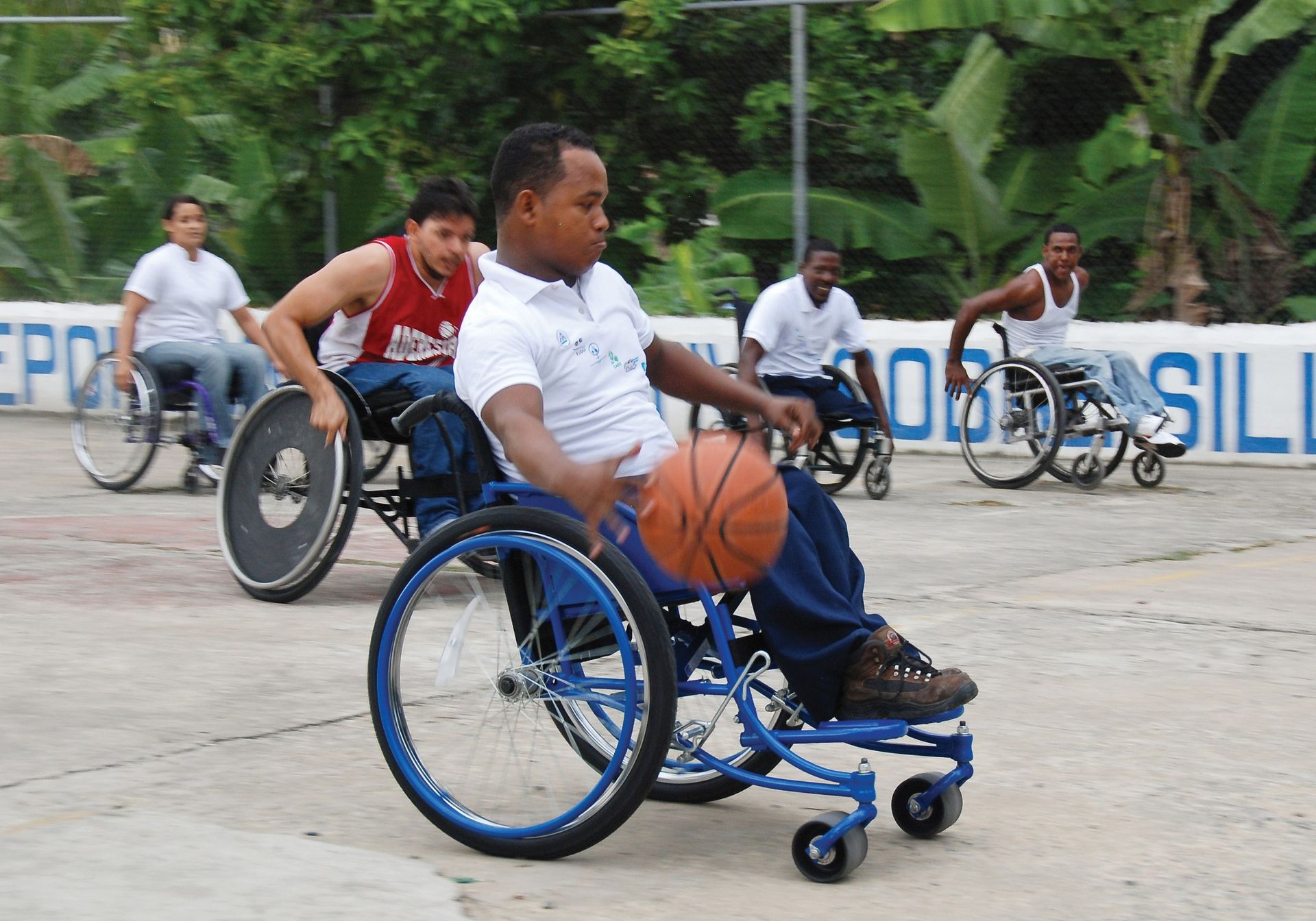 A man in a wheelchair bouncing a basketball with four men and one woman in wheelchairs on a basketball court.
