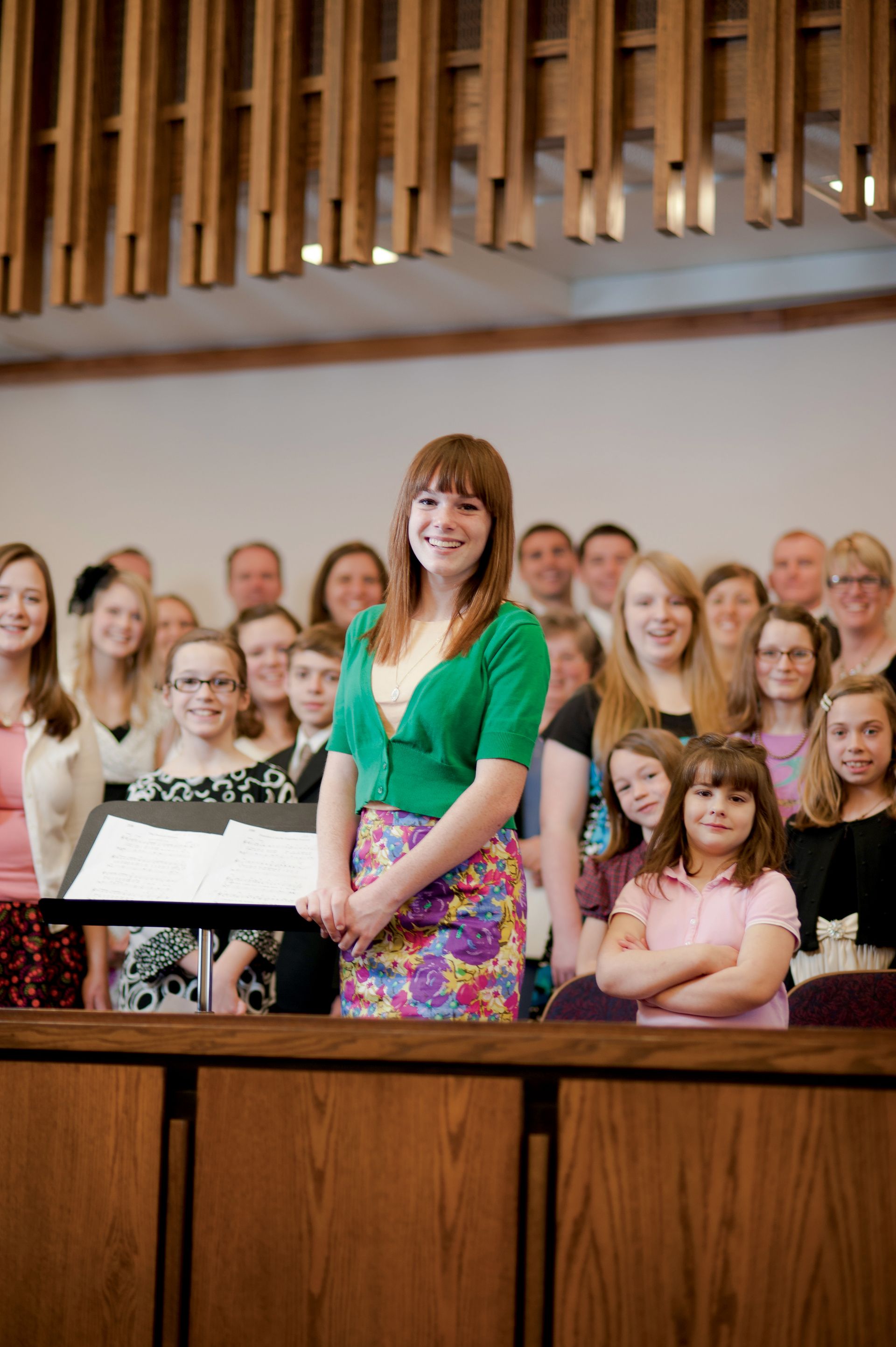 A young woman chorister stands in front of a choir of other young women.