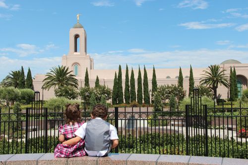 A little boy with brown curly hair putting his arm around his sister as they sit and look at the Newport Beach California Temple.