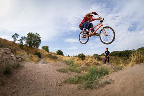 Two teenage boys go dirt biking, and one does a jump in the air while the other waits in the background.