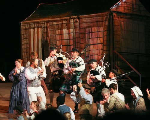 Nauvoo Pageant actors on a platform in front of a house, with four men playing bagpipes and other actors dancing.