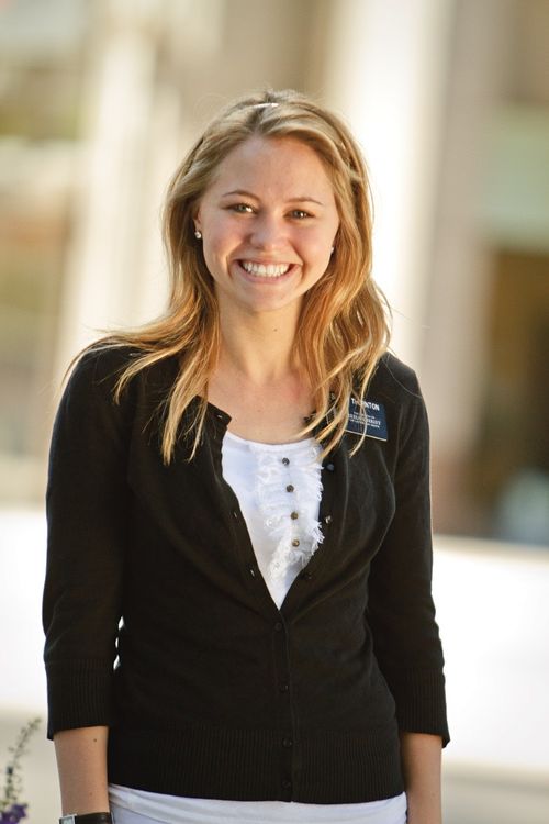 A portrait of a sister missionary with long blonde hair, a white shirt, and a black cardigan, standing and smiling.