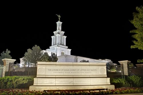 A sign reading “The Church of Jesus Christ of Latter-day Saints: Columbia River Washington Temple,” lit up at night with the temple in the background.