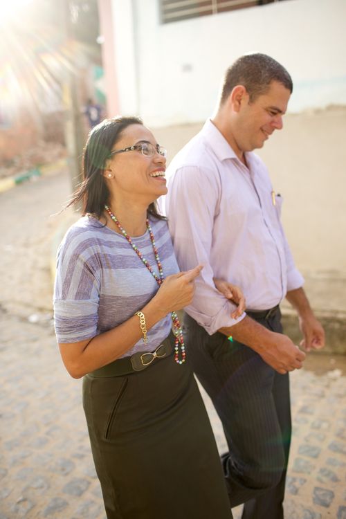 A husband in a button-up shirt and black pants laughs and walks arm-in-arm with his wife in a striped shirt and dark skirt.