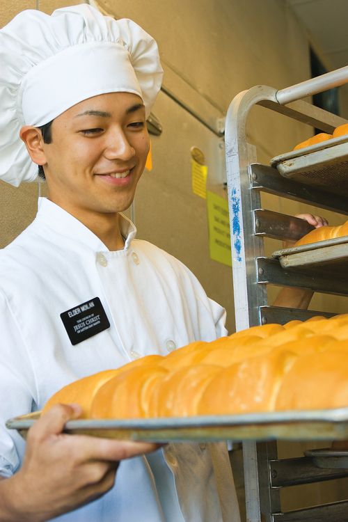 An elder missionary in a white apron and hat, serving in a bakery and carrying a pan with bread loaves.