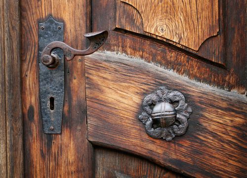 A old brown wooden door with two worn metal knobs fashioned in different shapes on the left side of the door.