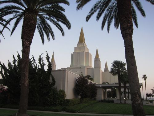 The Oakland California Temple and four of its spires seen between two palm trees that are growing on the temple grounds.