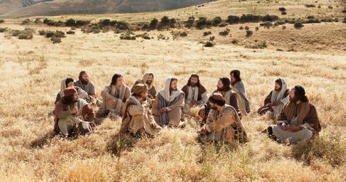 The disciples are gathered around Jesus, they are sitting together on the ground in a dry grassy field. One of the outtakes includes a lake in the background. Still from a Bible video. Jesus teaching a group in a field.