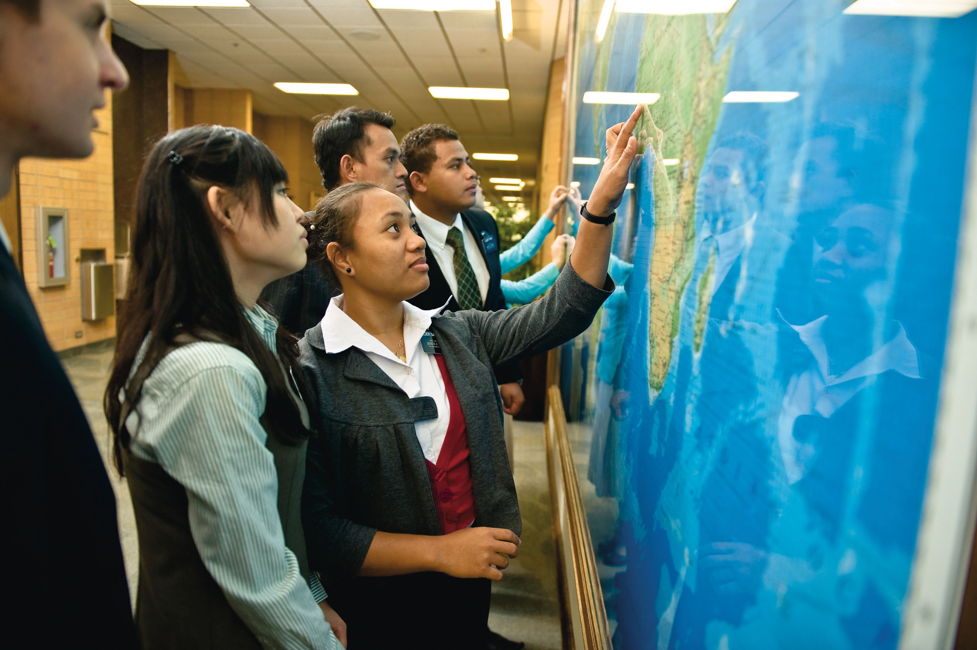 Elder and sister missionaries by a wall, pointing at a large map of the world.