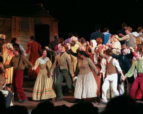 Actors dressed in pioneer clothing dancing together on a stage.