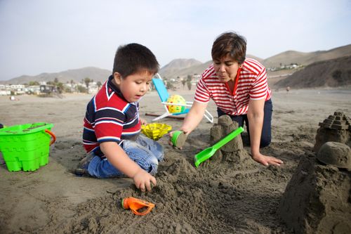 A grandmother kneels down in the sand next to her grandson and scoops up sand with him to build a sand castle.