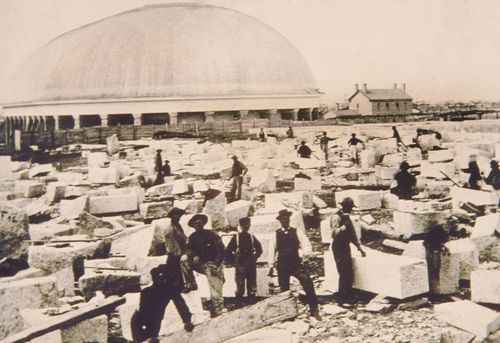 An old photograph showing workmen walking among large granite blocks on the piece of land next to the newly constructed Salt Lake Tabernacle.