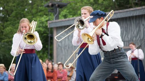 Two young female missionaries and a young male missionary playing trombones on an outdoor stage for an audience.