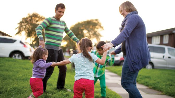 A man and woman playing “Ring around the Rosie” outside with children.