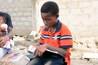 A young boy sits outside. He has scriptures with him. He appears to be studying. This is in Ghana, Africa.