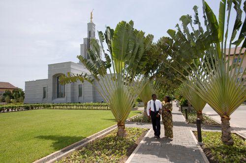 A couple walking with clasped arms down a sidewalk with the Accra Ghana Temple in the background.