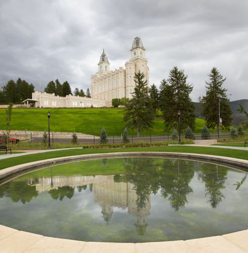 The Manti Utah Temple on a green hill with trees, seen reflected in the water of a pond on an overcast, rainy day.