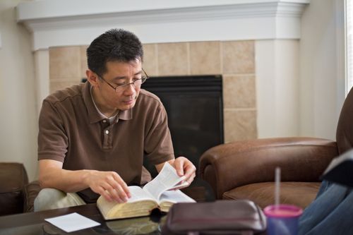 A man sits on a chair in his living room and flips the pages of a set of scriptures on the table in front of him.