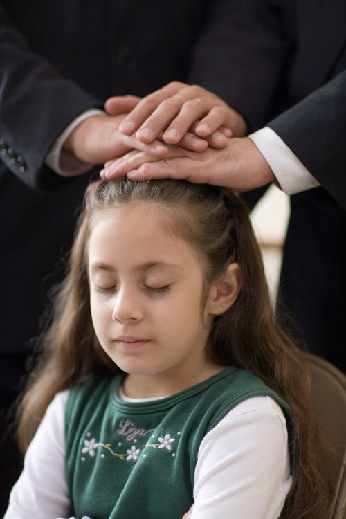 A girl with long brown hair bowing her head while two men in suits place their hands on her head to confirm her a member of the Church.