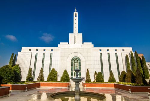 A side view of the Madrid Spain Temple, with a water fountain feature and stained-glass windows, on a sunny day with a blue sky behind.
