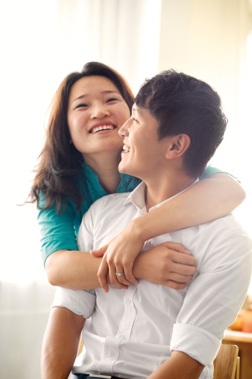 A portrait of a young Asian couple, with the man in a white shirt sitting down and his wife in a blue shirt standing and hugging him from behind.
