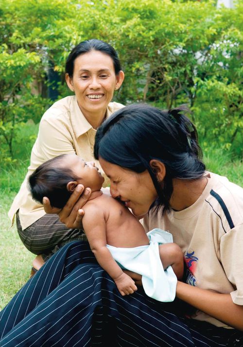A smiling Cambodian mother sitting outside and holding her baby close to her face, with another woman nearby.