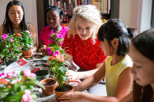 A group of girls and women gathered indoors planting flowers in small pots.