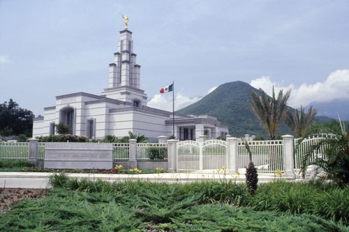 The Monterrey Mexico Temple on a sunny day, with green plants, trees, yellow flowers, the entrance gate and fences, the name sign in front, and a mountain seen in the distance.