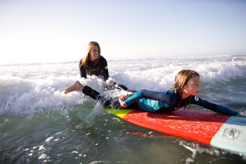 A mother sits on her surfboard in the water and watches her daughter paddle out in front of her on her surfboard.