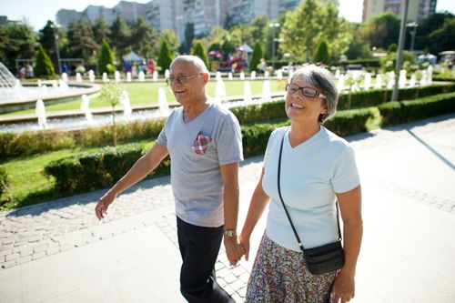 An elderly couple from Romania holding hands and walking down a sidewalk with the sun shining on their backs.