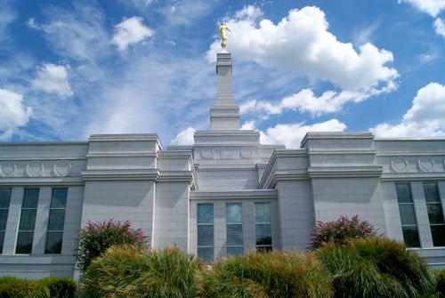 A view of the Palmyra New York Temple and spire from the ground, looking up toward a blue sky filled with white clouds.