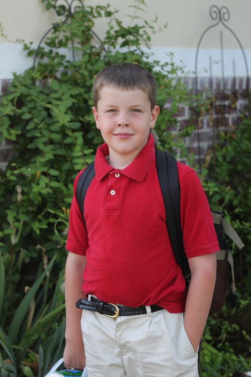 A portrait of a young boy wearing a red shirt, white pants, and a black backpack while standing outside.
