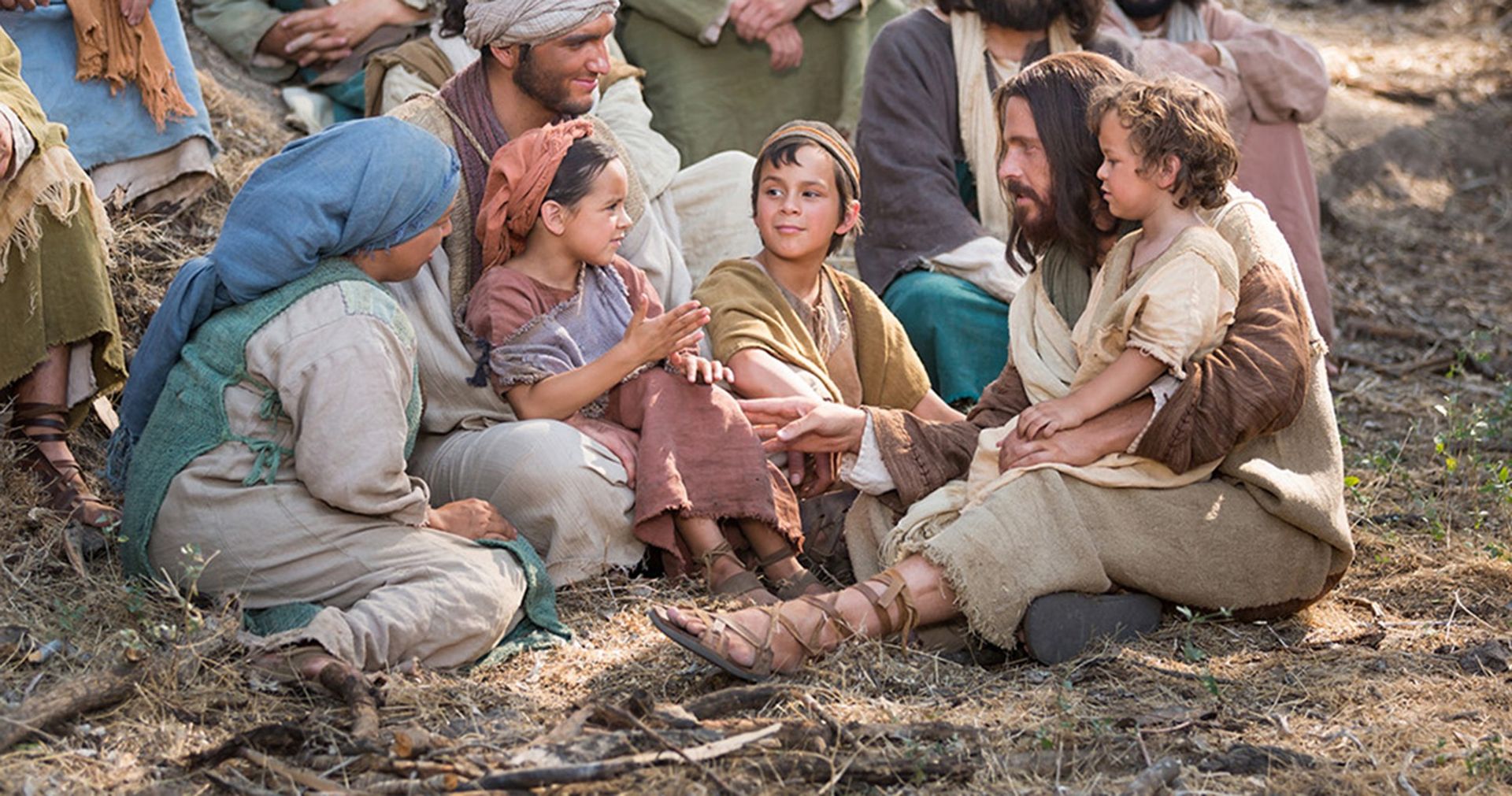 Jesus sitting on the ground with a young family, speaking directly to one child while holding another on His lap.