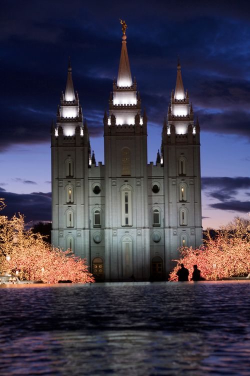 The front of the Salt Lake Temple lit up at night, with trees covered in Christmas lights on the temple grounds and a reflecting pond in the foreground.