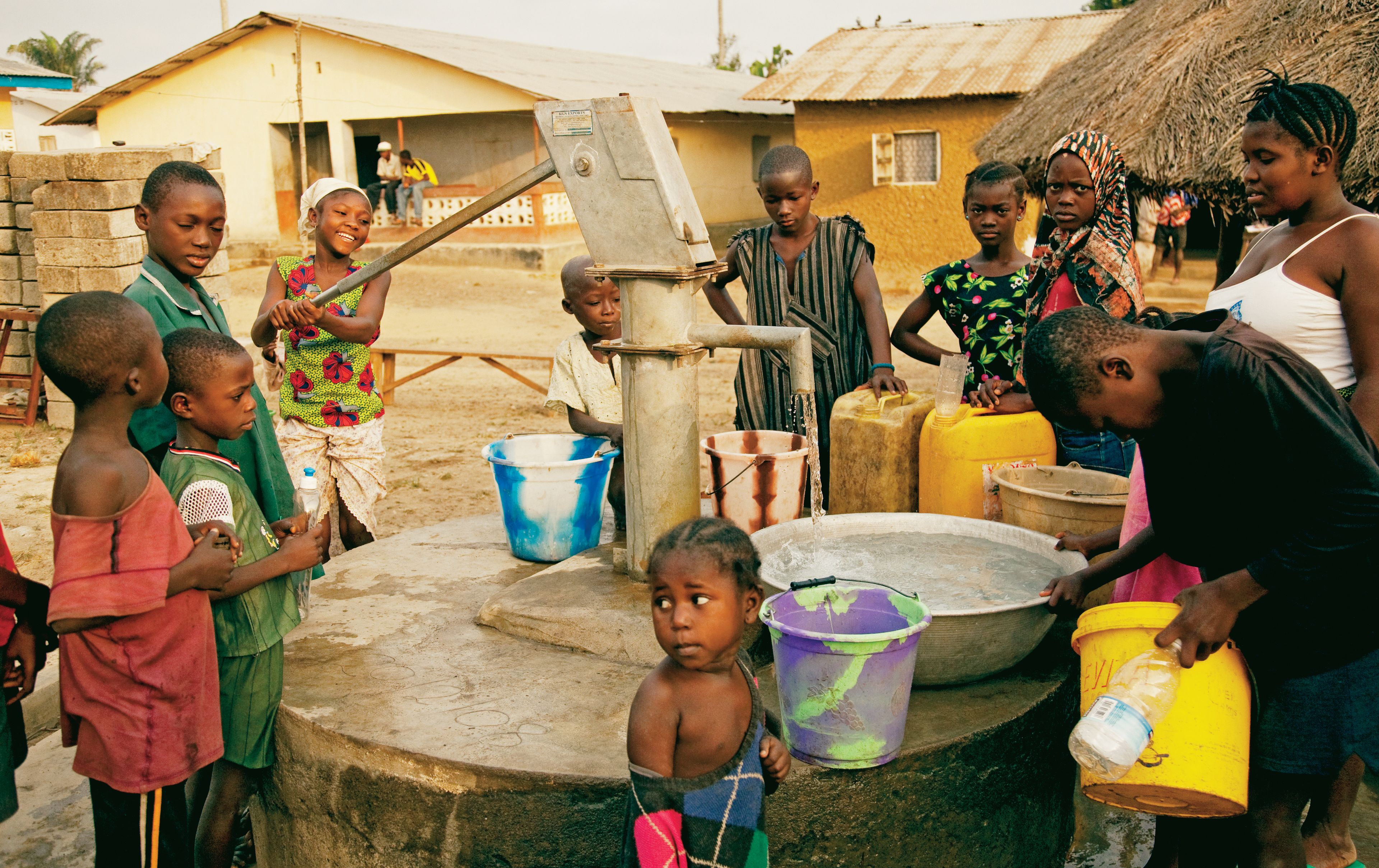African children and teenagers gather around with buckets while a young woman operates the water pump.