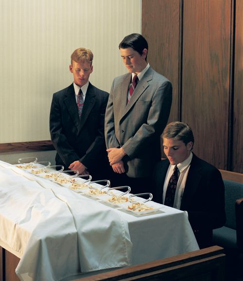 Two young men in suits stand near a sacrament table with their heads bowed while a third offers the prayer for the sacrament bread.