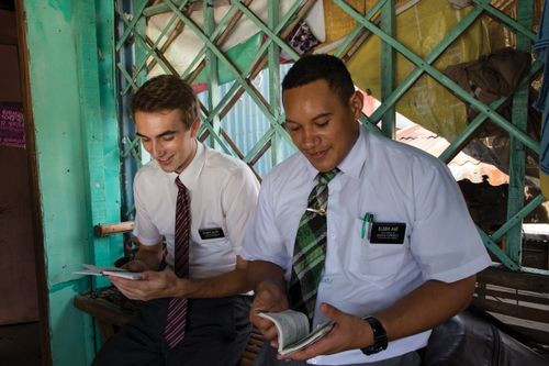 Two elder missionaries in white shirts sit inside a home and read aloud.