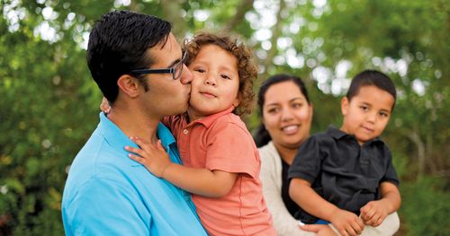 Young family in New Zealand playing and laughing with their children outside their home. They are playing basketball, holding the children and laughing and watching a small boy ride a trike.