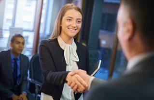 young woman steps forward as she is called in for her interview and shakes hands with her possible new employer. Behind her are some other candidates waiting for their turn .