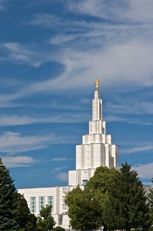 The top half of the Idaho Falls Idaho Temple rising above the trees on a sunny day, with a blue sky in the distance.