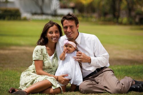 A husband and wife sit on the grass together with their baby daughter.