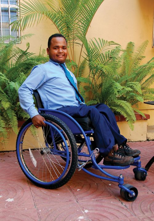 A man from the Dominican Republic, wearing blue pants, a blue shirt, and a blue tie, sitting and smiling in a new, shiny blue wheelchair.