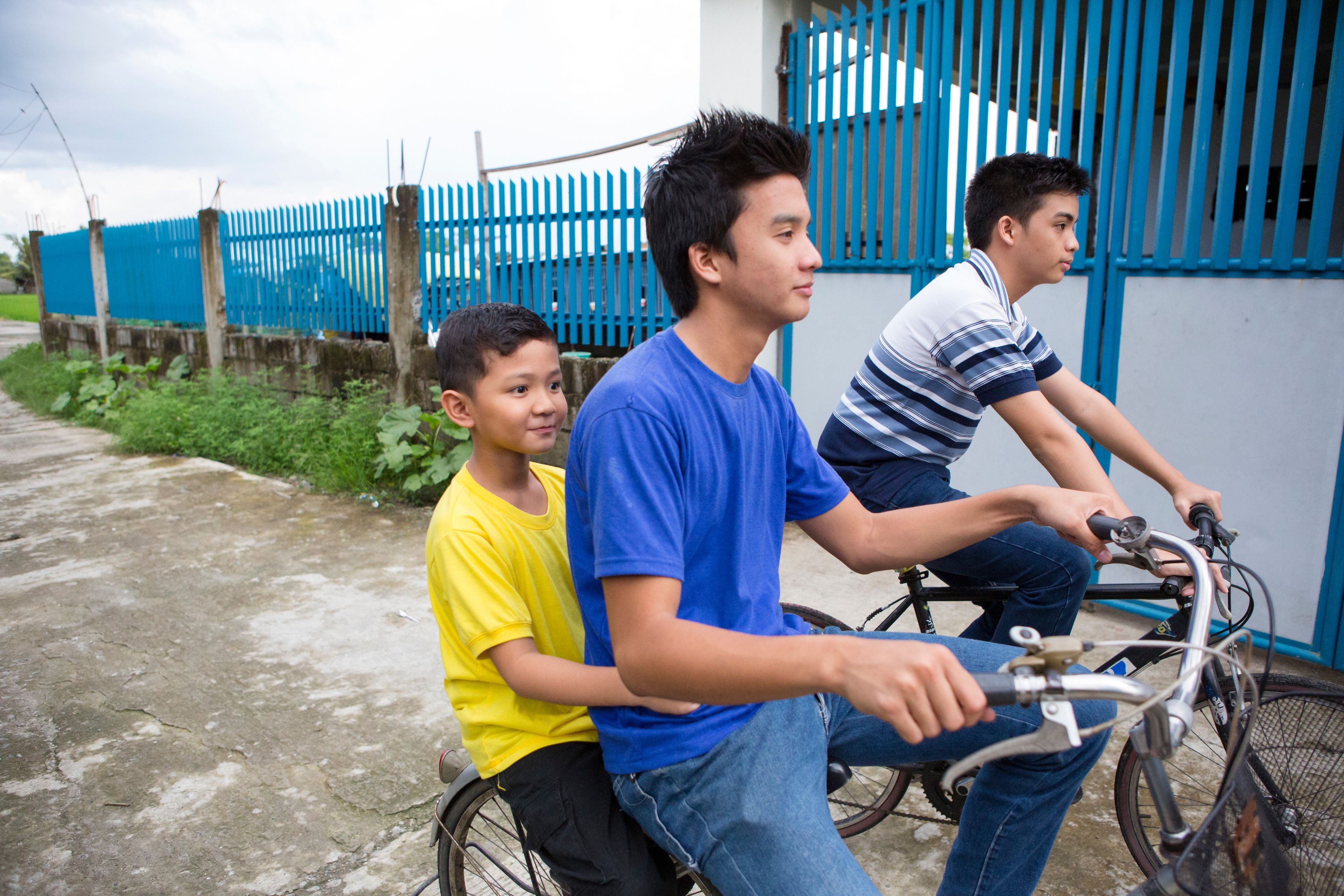 A young man rides a bike with a boy sitting behind him and another young man riding next to them.  