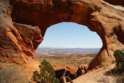 A large arch in the red rock mountains overlooking a valley and the clear blue sky.