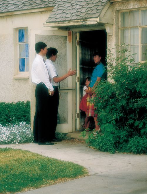 Two elder missionaries standing outside a home, talking to a woman in a blue shirt and her daughter in a red dress.