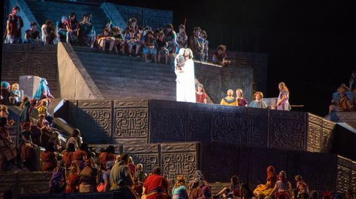 A large group of performers dressed in colorful clothing fill a large outdoor stage on a hillside.