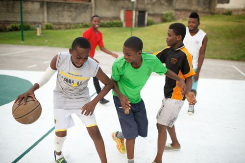 A young man plays with a group of other young men on an outdoor court in Africa, dribbling a basketball and guarding himself from another player.