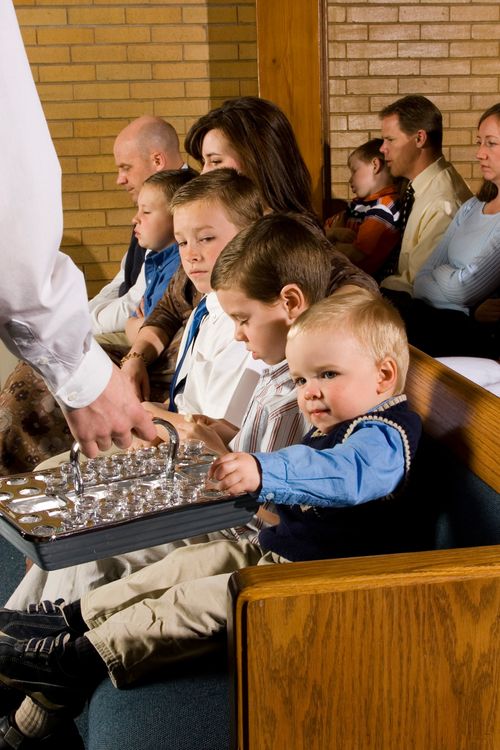 A blonde toddler reaching out to take a cup of water from a sacrament tray being passed to him in a sacrament meeting.