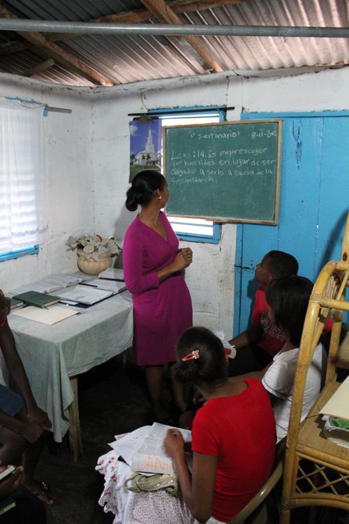 A woman in a dark purple dress stands in a small room next to a chalkboard and teaches seminary to three students, all sitting down in front of her.